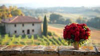 Red peony bouquet in ceramic vase on stone wall overlooking Tuscan countryside villa with cypress trees and rolling hills in soft evening light. Romantic rural scene.