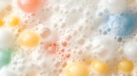 Colorful soap bubbles and foam macro photography with yellow, coral pink, and turquoise tones creating abstract pattern on white background. Soft dreamy texture.