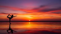 Woman practicing yoga pose on beach at sunset with dramatic colorful sky reflecting in calm water creating peaceful meditation scene for wellness lifestyle.