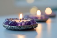 Glowing candle nestled in raw amethyst crystal holder creates serene atmosphere with soft bokeh lights in background, spiritual wellness concept.