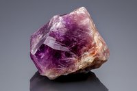Raw amethyst crystal specimen displaying vibrant purple and orange hues with natural geometric formations against dark background, showcasing mineral's rough texture and translucency.