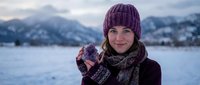 Young woman in purple knit hat and mittens holding hot drink in snowy mountain landscape during winter vacation enjoying warm beverage outdoors.