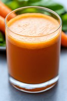 Fresh carrot juice in clear glass with blurred carrots and green leaves in background. Bright orange healthy drink with natural foam on top. Macro food photography.