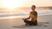 Young woman practicing yoga meditation on beach at golden hour sunset with peaceful ocean waves in background creating serene wellness lifestyle moment.