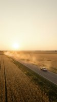 Vehicle traveling on rural highway through golden wheat fields at sunset, creating dust clouds across expansive agricultural landscape for travel and farming concepts.