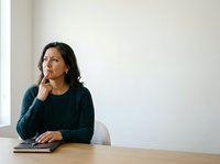 Middle-aged woman of South Asian descent sitting at wooden desk wearing dark teal sweater, hand to chin in thoughtful pose, looking upward contemplatively with notebook nearby, suitable for business, education, and professional concepts.
