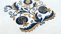 Ornate blue and gold floral embroidery design on white fabric featuring decorative swirls and botanical motifs for textile crafts and needlework projects.