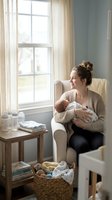 Young mother breastfeeding newborn baby in cozy nursery chair by window with natural light and feeding supplies on wooden table for motherhood care.