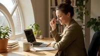 Young Caucasian businesswoman smiling while working on laptop at wooden desk in bright home office with plants and books.