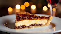 Slice of layered chocolate pecan pie with creamy custard filling on white plate against warm bokeh lights background, close-up view of homemade dessert.