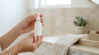Caucasian woman's hands holding white cosmetic bottle in bright bathroom with natural light, displaying skincare product for beauty and wellness marketing.