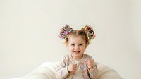 Young girl with blonde hair, approximately 3-5 years old, wearing colorful hair bows, smiling at camera against neutral background. Suitable for children's products, parenting content, and family-oriented designs.