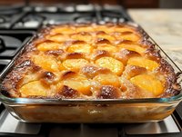 Fresh peach cobbler with golden brown crust baked in glass baking dish on stovetop, showing caramelized fruit and buttery lattice top, homestyle dessert.
