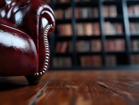 Luxurious burgundy leather armchair detail with decorative nailhead trim against blurred library bookshelves background on wooden floor. Cozy reading atmosphere.