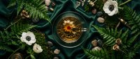 Blooming flower tea in a glass cup surrounded by ferns, anemone flowers, stones, and moss on dark green silk fabric, flat lay.