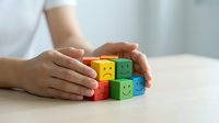 Hands protecting colorful wooden blocks with emoji faces showing mental health support and emotional wellbeing care in workplace or therapy setting.