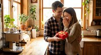 Happy couple sharing romantic moment with red gift box in cozy kitchen during golden hour sunlight for Valentine's Day celebration.