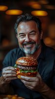 Mature Caucasian man with gray beard smiling while holding fresh juicy burger in restaurant. Dark moody atmosphere with bokeh lights creates cozy ambiance for food photography.