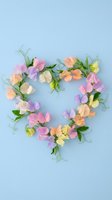 Heart-shaped wreath made of colorful sweet pea flowers with green leaves and tendrils arranged on soft blue background for romantic decoration.
