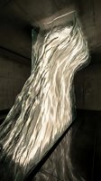 Glowing glass sculpture with flowing wave texture stands in a dark concrete interior, illuminated from within, creating dramatic light and shadow.