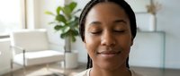Young Black woman with braided hair smiling with eyes closed in bright modern interior with plant and white furniture, expressing contentment and relaxation.