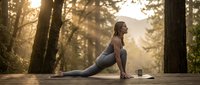 Young woman practicing yoga lunge pose on wooden deck in forest at golden hour sunrise, peaceful morning meditation and fitness routine outdoors.