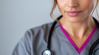 Close up portrait of healthcare worker in gray scrubs and pink undershirt with stethoscope, showing confident smile against light background. Professional medical staff concept.