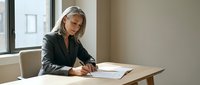 Mature businesswoman with gray hair writing documents at office desk near window in modern workplace setting for professional corporate content.
