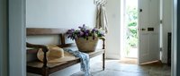 Rustic wooden bench with wicker basket of purple flowers and folded blanket in bright entryway interior with natural light from open door.