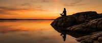 Woman sitting on rocky outcrop watching golden sunset over calm water, silhouetted against orange sky. Peaceful contemplation scene.