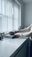 Woman in cozy sweater reading book with tea cup by window with sheer curtains in minimalist modern interior for relaxation and lifestyle content.