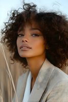 Young African American woman with natural curly hair and glowing skin wearing beige blazer, looking up with confident expression against soft natural background.