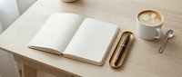 Open notebook with blank pages, gold pen, and cappuccino on wooden desk for creative writing, planning, and workspace design projects.