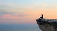 Silhouette of person sitting alone on cliff edge against pastel sunset sky and misty mountain landscape, contemplative solitary moment in nature.