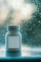 White medicine bottle with blank label against rainy window background, creating moody atmosphere with water drops and soft teal bokeh effect.