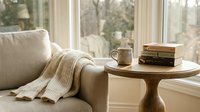 Cozy reading nook with beige armchair, knitted throw blanket, coffee mug and books on wooden side table by large windows with natural light.
