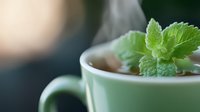 Steaming hot herbal tea in mint green ceramic cup decorated with fresh mint leaves, close-up view with soft natural lighting and shallow depth of field.