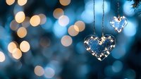 Glass heart ornament with fairy lights against blurred blue and gold bokeh background creates romantic holiday atmosphere for Valentine's Day or Christmas decor.