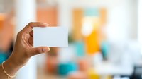 Person with dark skin tone holding blank white business card in office environment with colorful blurred background. Use for corporate branding, networking, and professional presentations.