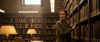 Young professional woman browsing books in traditional library with warm lighting and wooden shelves creating scholarly atmosphere for research.