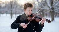 Young Caucasian male violinist performing classical music outdoors in snowy winter park with falling snowflakes creating magical atmosphere for concerts.