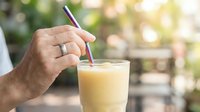 Man's hand holding striped straw in glass of pale yellow beverage outdoors with blurred garden background, suitable for summer drink, lifestyle, and refreshment concepts.