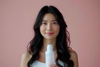 Young Asian woman with long dark hair holding white cosmetic spray bottle against soft pink background for skincare and beauty product advertising.