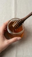 Hand holding glass jar of golden honey with wooden dipper dripping sweet natural syrup on neutral linen background for organic food and wellness concepts.
