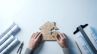 Real estate agent assembling wooden house puzzle pieces on desk with architectural blueprints and plans for property development and construction projects.