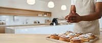 Young male baker with medium skin tone presenting freshly baked pastries on wooden board in modern kitchen, wearing white apron. Ideal for bakery, culinary, and food business marketing materials.