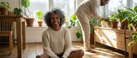 Smiling mature African American woman sitting on floor at home while man tends to houseplants in bright living room with wooden decor.