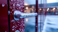 Frozen door handle covered in ice crystals and frost on red metal door during winter storm, macro close up view with blurred background and bokeh lights.
