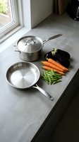 Stainless steel cookware and fresh vegetables arranged on gray kitchen counter near window, ready for meal preparation and cooking.