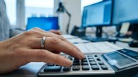 Professional businessman calculating finances using calculator at modern office desk with dual computer monitors for accounting and budget planning tasks.